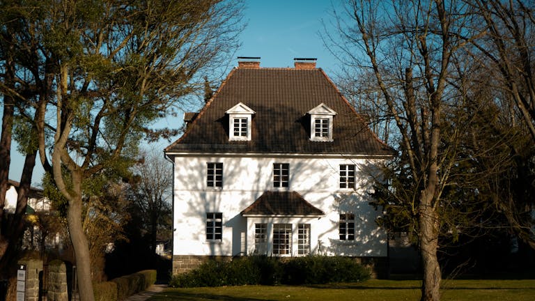 Elegant colonial-style house with traditional roof amid lush trees on a sunny day.